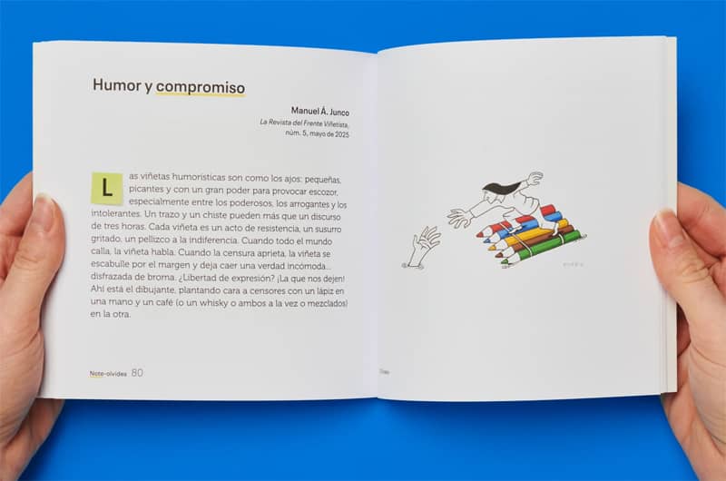 Fotografía del libro abierto sostenido por dos manos sobre un fondo azul. En la página de la izquierda, bajo el título "Humor y compromiso", aparece un texto de Manuel Á. Junco que reflexiona sobre el poder de la viñeta como acto de resistencia. En la página de la derecha, se muestra una ilustración de Eneko de una persona navegando sobre un conjunto de lápices de colores atados como una balsa, estirando el brazo para rescatar a otra persona que se hunde en el agua.